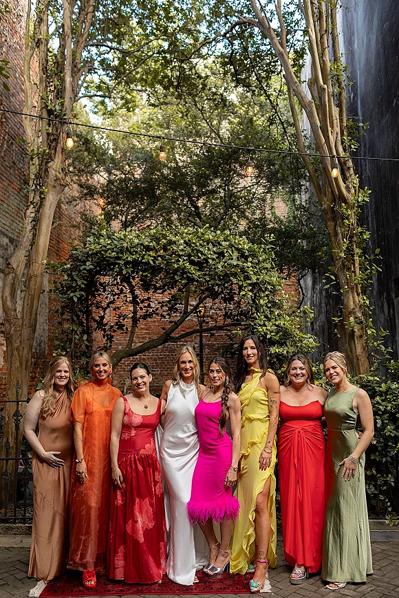 Bridal party portrait of bride with bridesmaids in colorful bridesmaid dresses, posed on a stone walkway in a garden courtyard under string lights