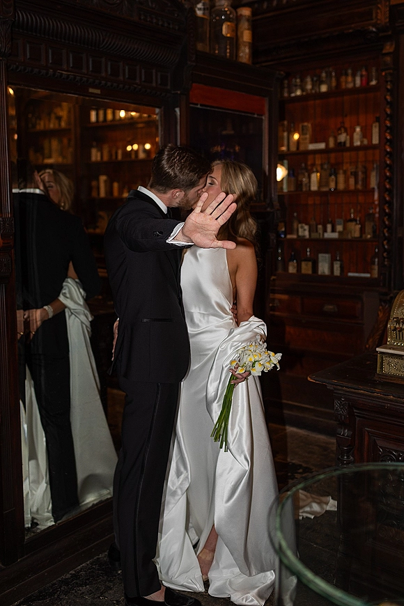 Wedding kiss as bride in satin dress holds a calla lily bouquet while groom in black tux kisses in a moody bar interior