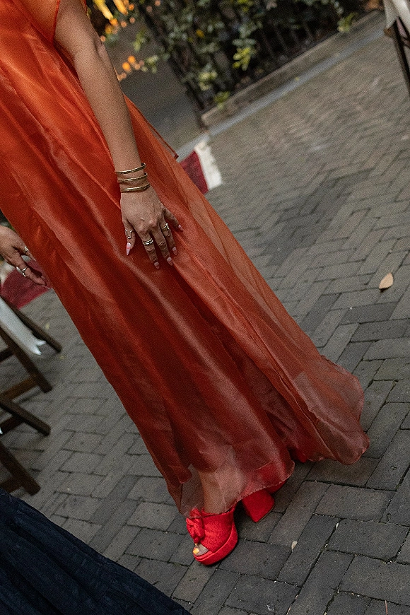 Wedding guest outfit featuring an orange chiffon dress with red high heels, stacked bangles, rings, and manicure on a tree-lined walkway