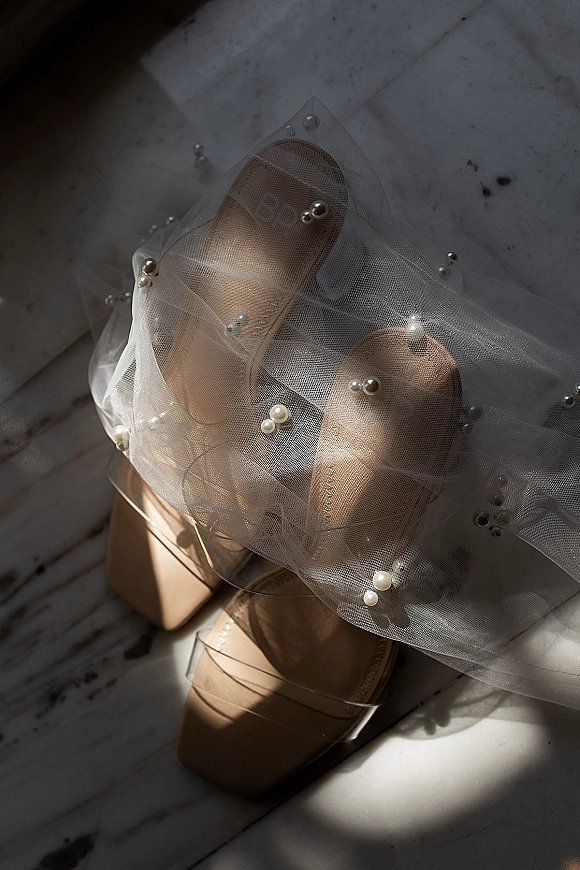 Bridal shoes set in a wedding shoes flatlay with pearl embellishments on a white marble floor, lit by window light and soft shadows