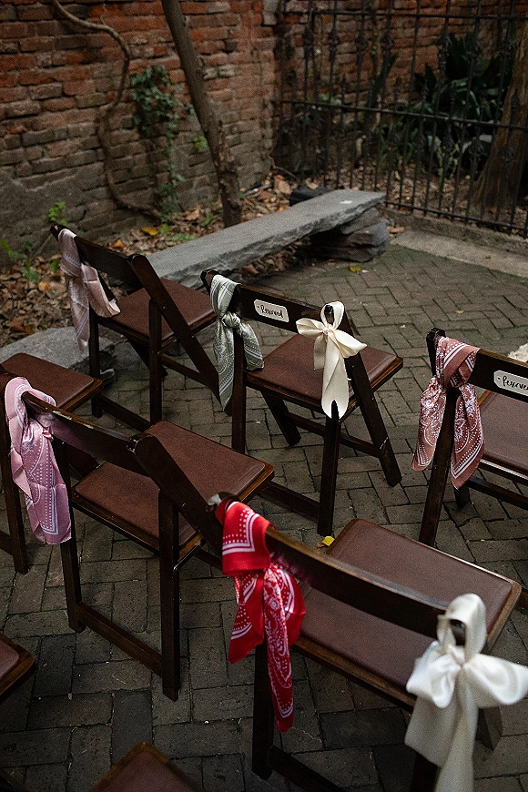Ceremony seating with wedding ceremony chairs, wood folding chairs tied with ribbons and bandanas beside a brick courtyard wall and iron fence
