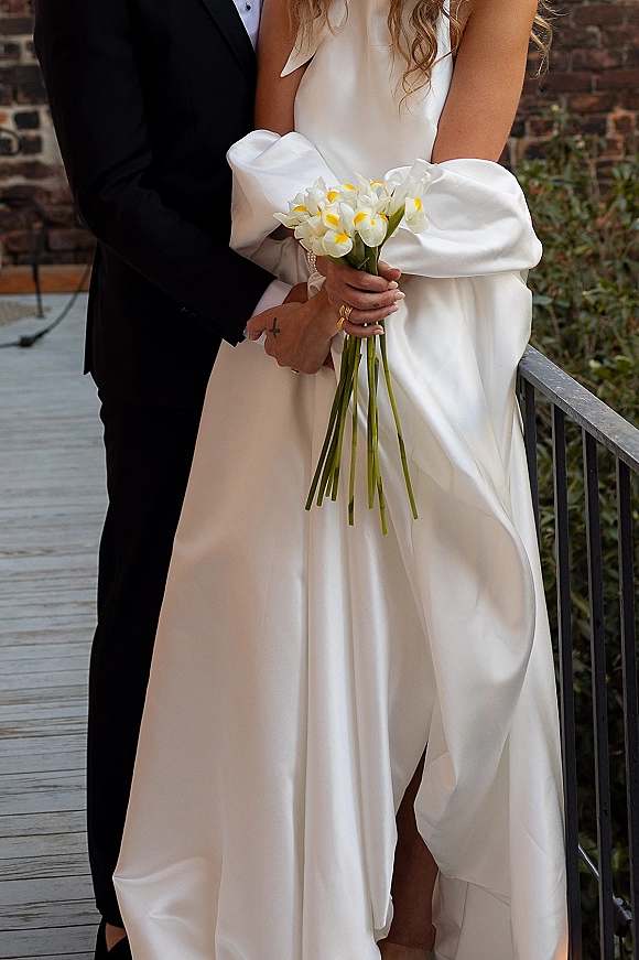 Couple portrait of bride and groom embrace, her calla lily bouquet against a brick wall backdrop beside a metal railing
