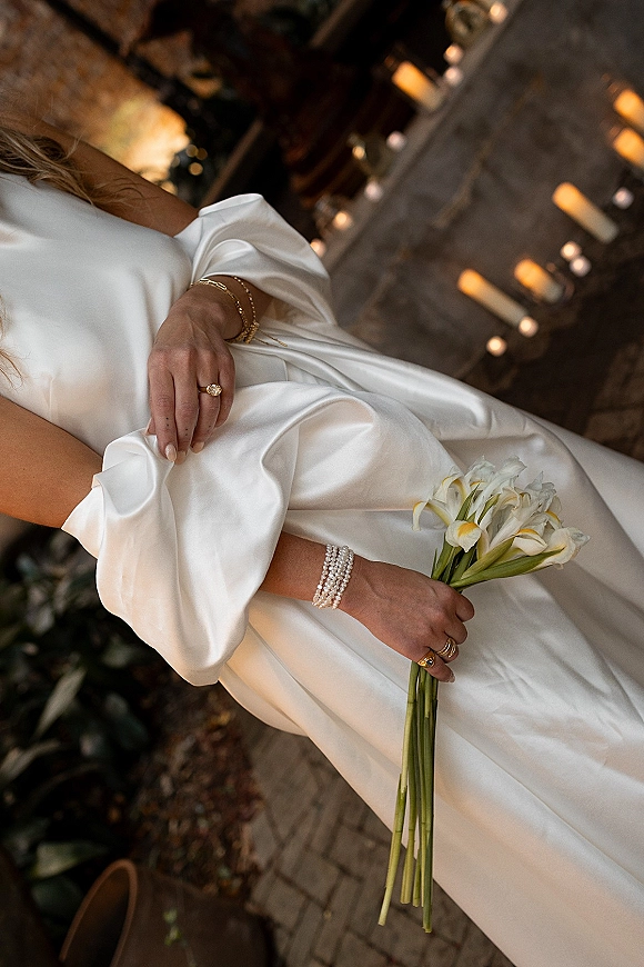 Bridal portrait with calla lily bridal bouquet held close, showing satin wedding dress and gold jewelry by candlelit stone fireplace.