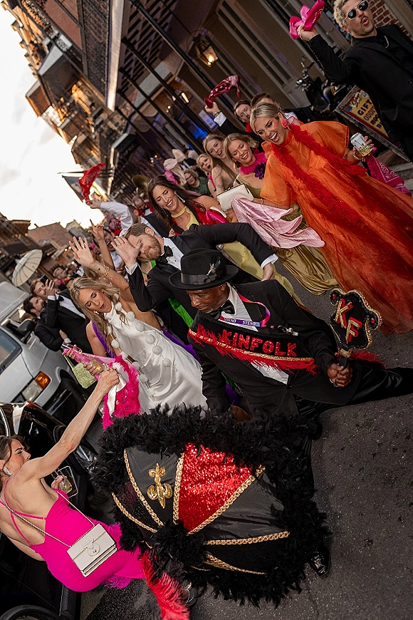 Wedding second line parade with bride in white gown and groom in tuxedo leading guests waving handkerchiefs on a dusk city street