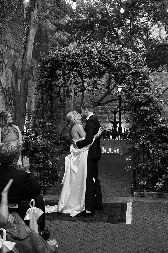 Wedding kiss under a floral arch, bride in off-the-shoulder gown and groom in black tux amid string lights and candles in a brick courtyard