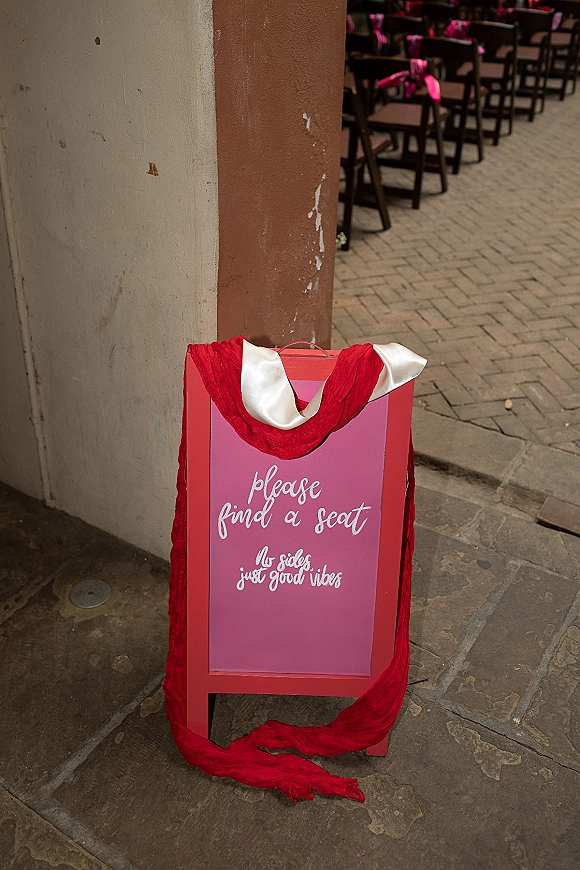 Wedding signage with a please find a seat sign on an A-frame, hand-lettered and draped with red fabric in a courtyard aisle entrance