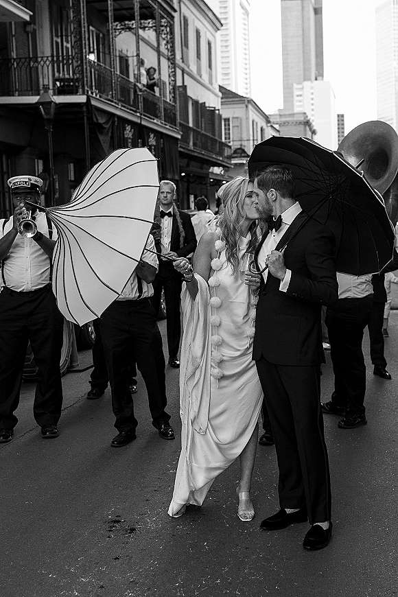 Wedding kiss portrait of newlyweds kissing under umbrellas, bride in gown and groom in tuxedo on a city street with brass band behind