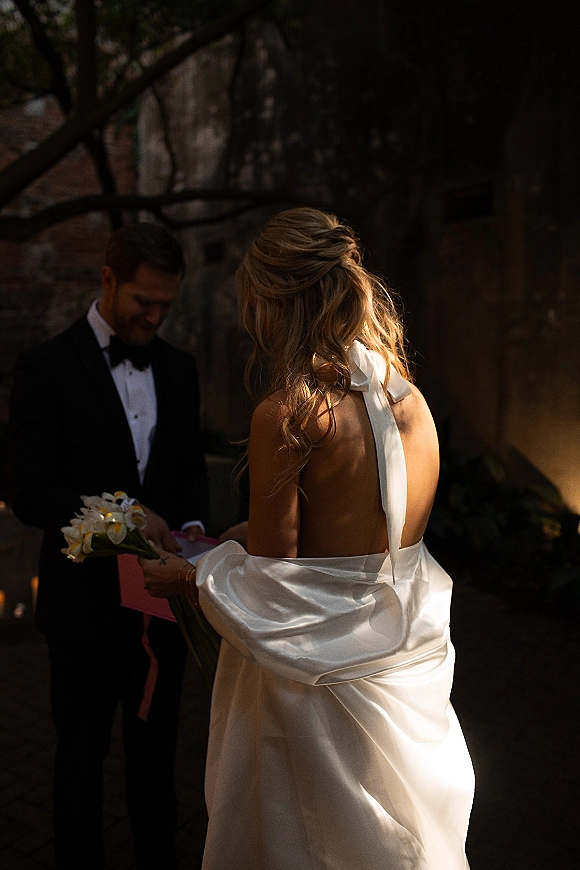 Couple portrait of bride in a backless wedding dress with bow detail holding a calla lily bouquet beside groom in black tuxedo outdoors by stone wall