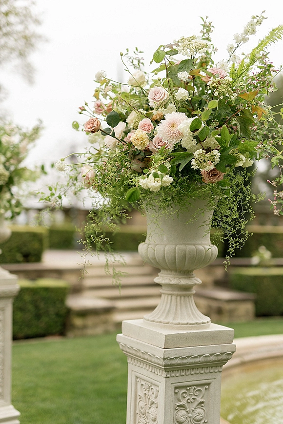 Wedding floral arrangement of blush roses, dahlias, and hydrangea overflowing from an urn vase on a pedestal by stone steps in a garden