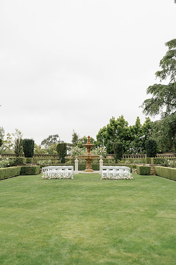Outdoor ceremony setup with white ceremony chairs in rows and floral aisle arrangements facing a stone fountain on a manicured garden lawn