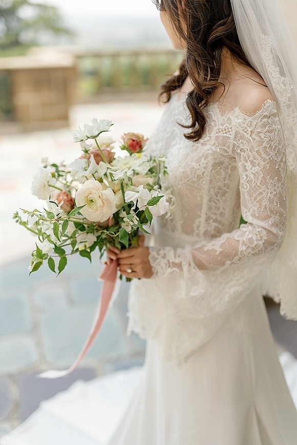 Bridal portrait of a bride holding bouquet in side profile, wearing a long sleeve lace dress and veil on an outdoor terrace with greenery.