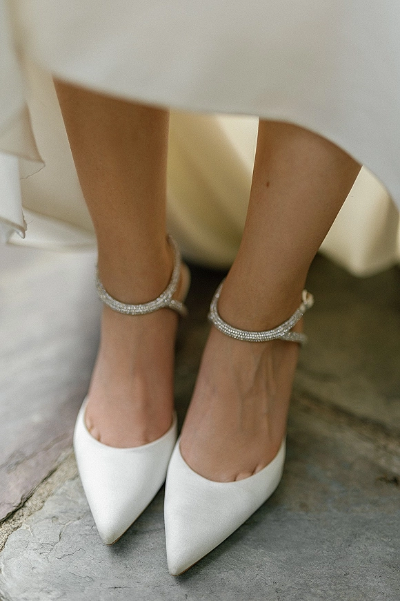 Bridal shoes with white wedding heels and rhinestone ankle straps beneath the wedding dress hem on an indoor stone floor