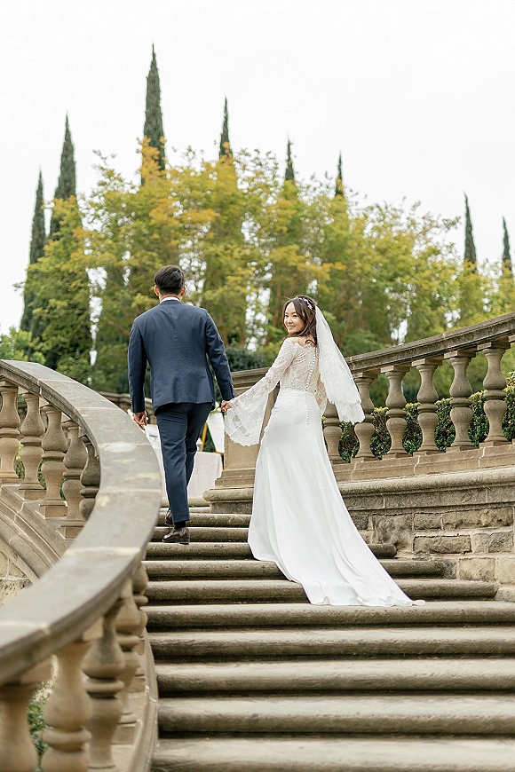 Couple portrait of bride and groom holding hands on stone staircase, her long veil and lace sleeves flowing as she looks back