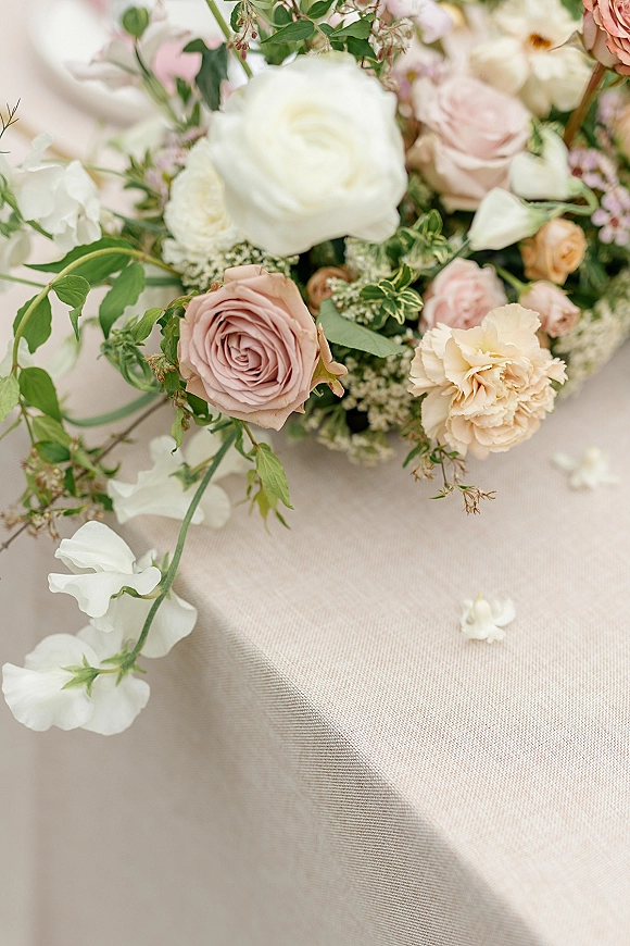Wedding floral centerpiece with blush and white roses, ranunculus, carnations, and greenery arranged low on a neutral tabletop surface