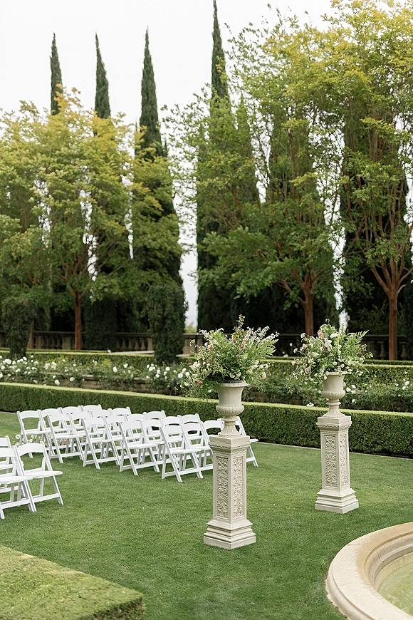 Ceremony setup with white folding chairs and floral urns on pedestals lining the aisle, set on a manicured lawn by a fountain