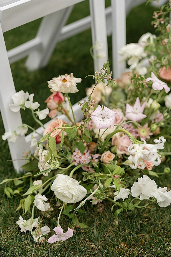 Wedding aisle florals with ceremony chair flowers in a pastel wildflower meadow of roses, ranunculus, and greenery on a grass lawn