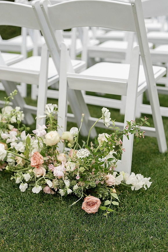 Ceremony aisle flowers with a ground floral arrangement of roses, sweet peas, and greenery lining white folding chairs on a grass lawn