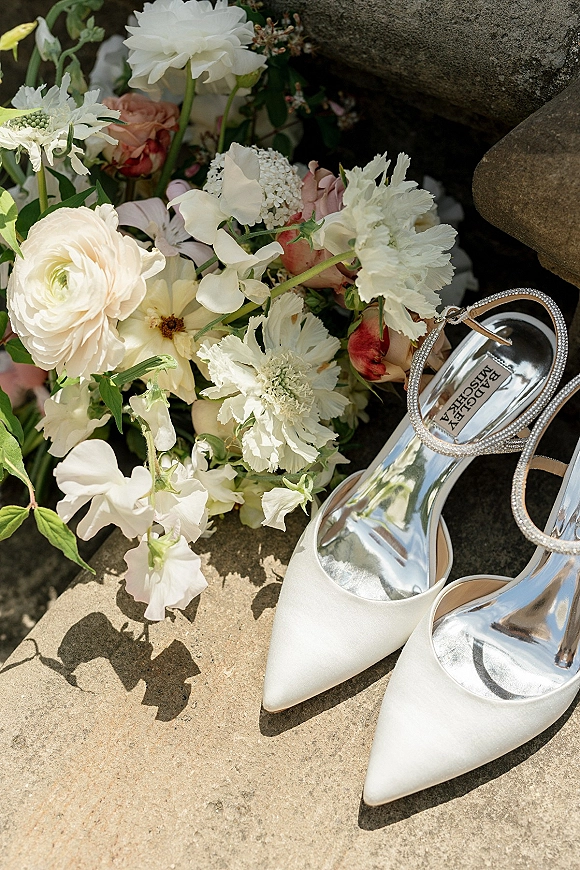 Wedding shoes flatlay with white bridal heels and rhinestone ankle straps beside a blush rose bouquet on a stone surface