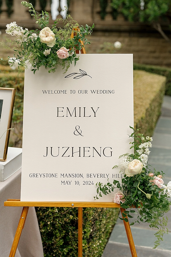 Wedding welcome sign on a wooden easel with blush and white rose accents beside a framed photo, set along garden stone steps and hedges
