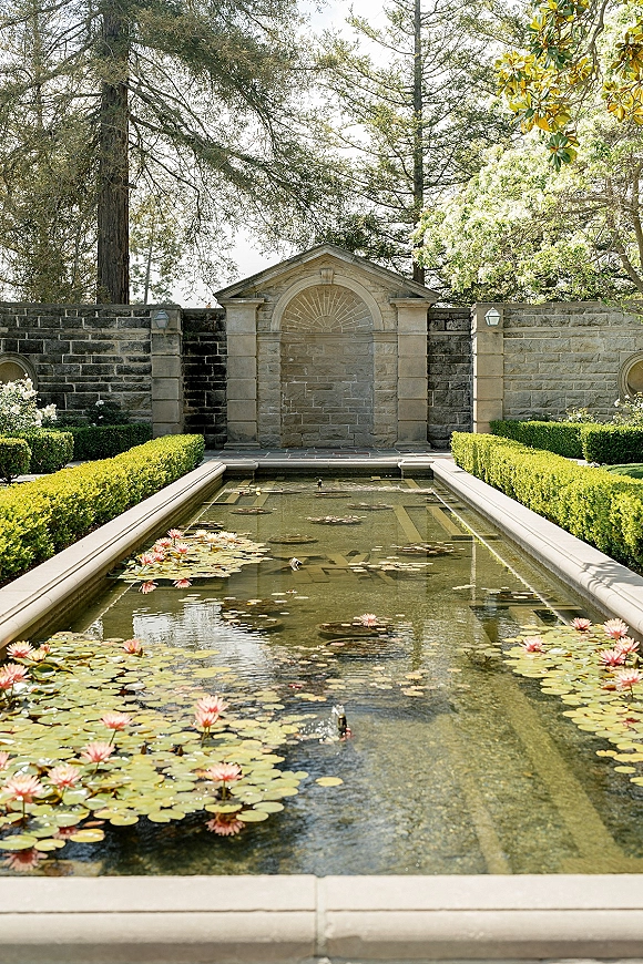Garden wedding venue with a reflecting pool filled with water lilies, trimmed hedges, and a stone fountain wall beneath tall trees and garden lights
