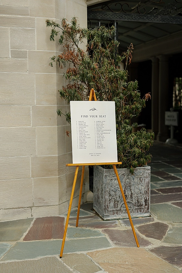 Wedding seating chart find your seat sign on a wooden easel with a potted plant, set against a stone wall in a covered walkway