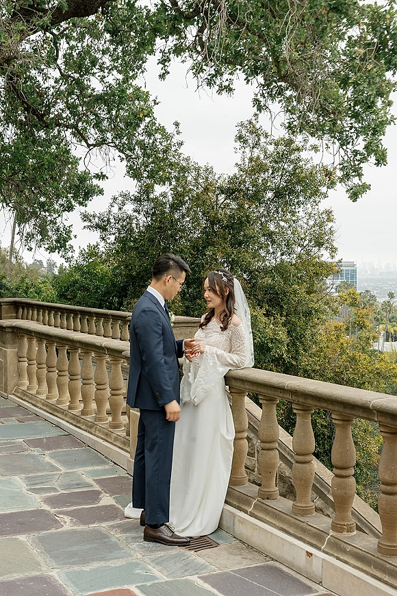 Couple portrait of bride in lace long-sleeve gown and veil holding hands with groom in navy suit on a stone terrace railing outdoors