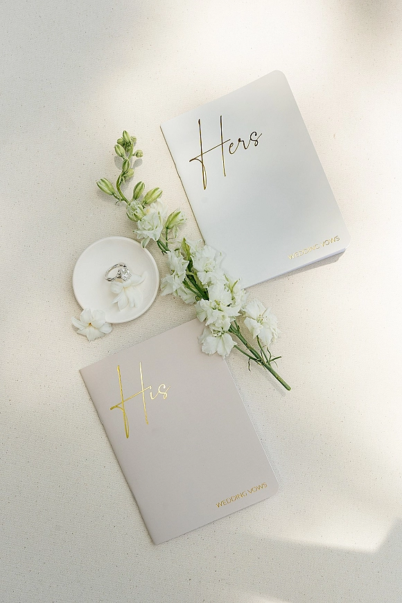 Wedding vow books with gold foil lettering beside a ring in a small dish and white flowers on neutral linen fabric