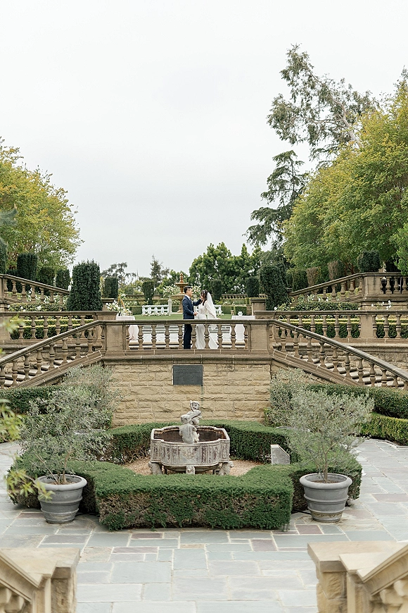 Wedding couple portrait of bride and groom in garden kissing on a stone terrace by a fountain, bridal veil flowing behind them