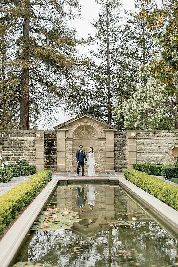 Couple portrait of bride and groom holding hands, bride with bouquet in long-sleeve gown beside a reflecting pool with lily pads in a garden courtyard