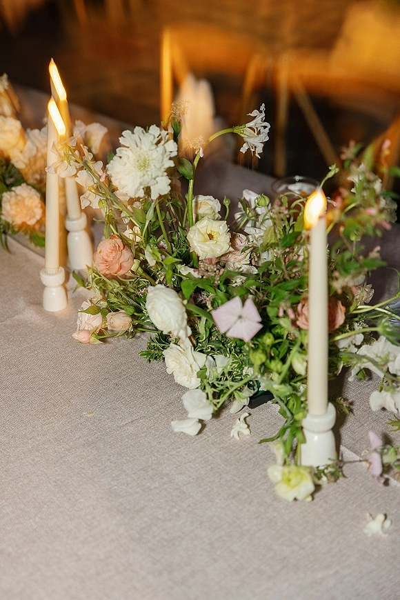 Wedding centerpiece with garden style centerpiece florals, white taper candles in ivory holders, greenery on linen table in dim reception light