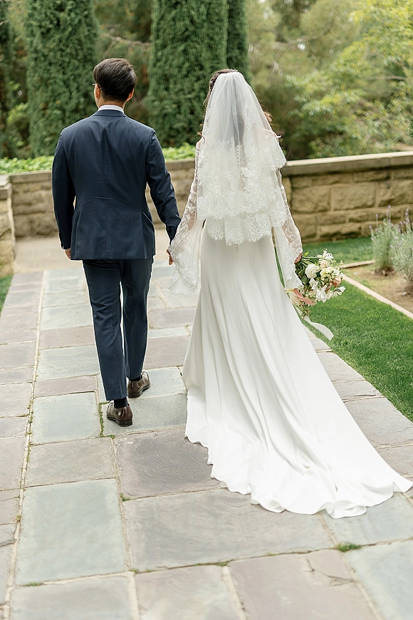 Couple portrait of bride and groom walking away holding hands, her lace veil and gown train flowing on a stone garden walkway