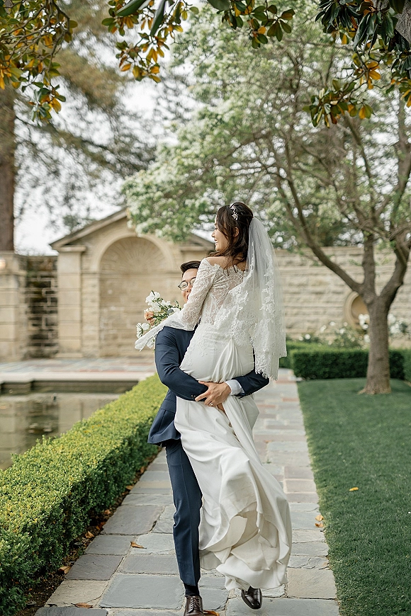 Couple portrait of groom holding bride in his arms, her long lace veil flowing as they smile by a reflecting pool in gardens