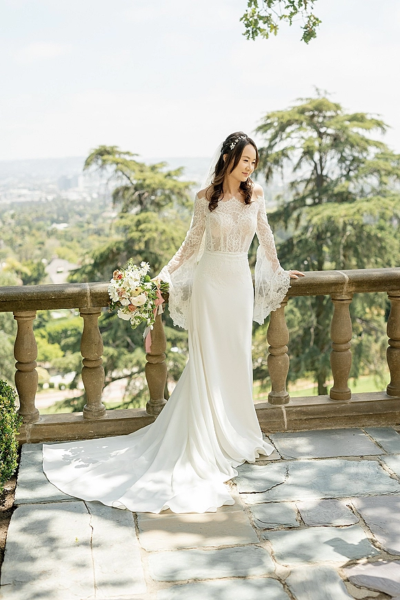 Bridal portrait of a bride leaning on a stone terrace railing, holding a bouquet with a pink ribbon, in a lace long sleeve dress and cathedral veil