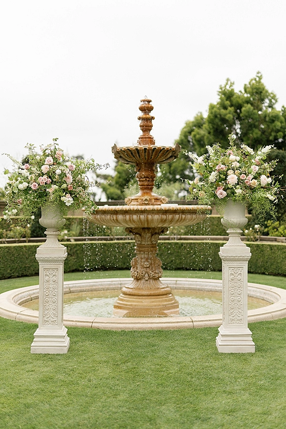 Wedding ceremony backdrop with a stone fountain and rose-and-greenery urn florals, framed by carved columns on a manicured garden lawn