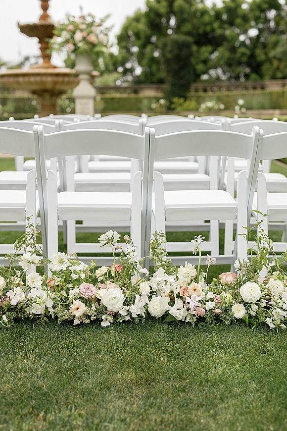 Ceremony seating with white wedding chairs lined on a lawn aisle, bordered by low rose and ranunculus florals with greenery near a stone fountain