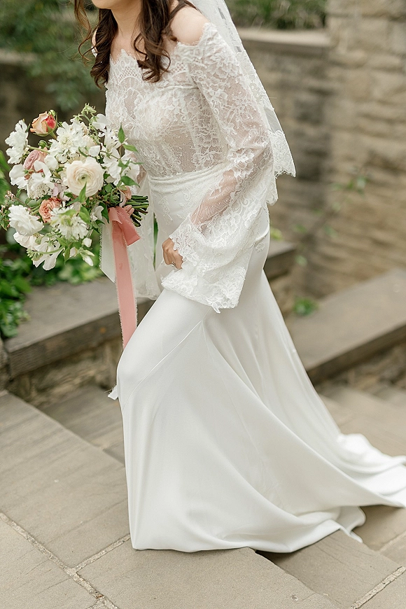 Bridal portrait of a bride in a lace sleeve wedding dress holding a rose bouquet with blush ribbon on stone steps by garden greenery