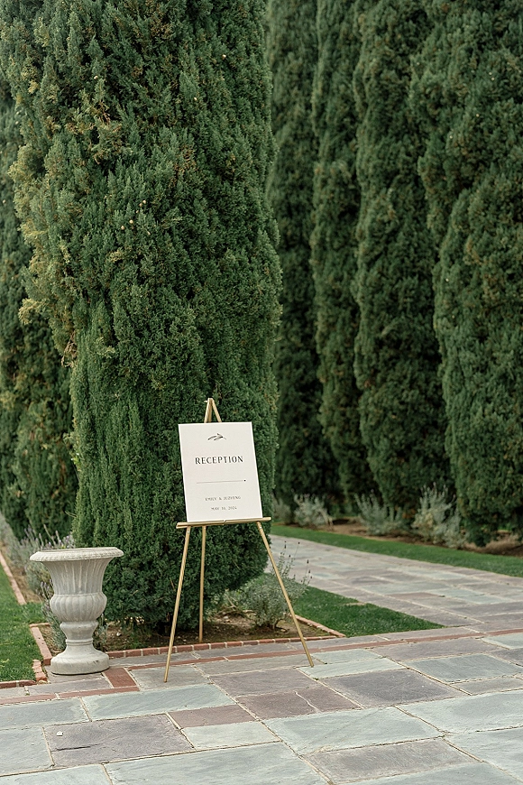 Wedding reception sign on a gold easel with white cardstock welcome text beside a stone urn along a garden walkway with cypress trees