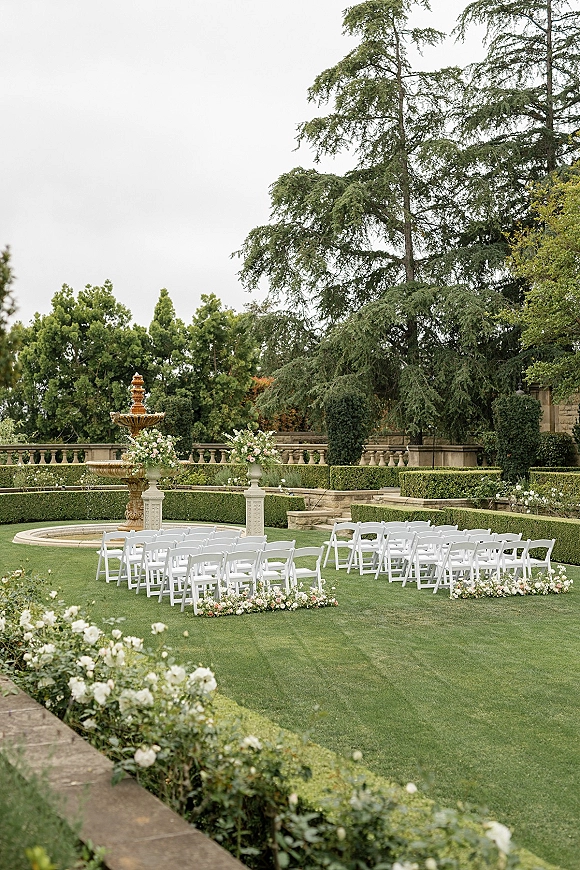 Outdoor ceremony setup with garden wedding ceremony seating in a semicircle of white folding chairs, floral aisle markers, and a stone fountain on a manicured lawn