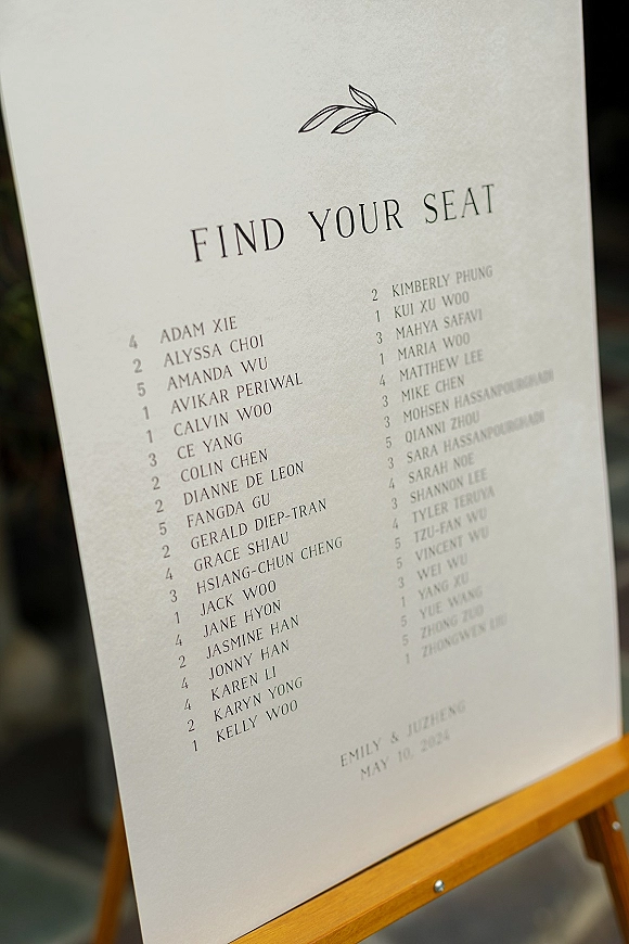 Wedding seating chart with a find your seat sign printed on a poster and displayed on an easel along a greenery-lined outdoor walkway
