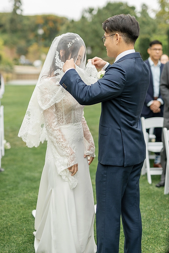 Ceremony moment as groom lifting veil from bride in long-sleeve lace dress, guests seated on white chairs on a grassy lawn