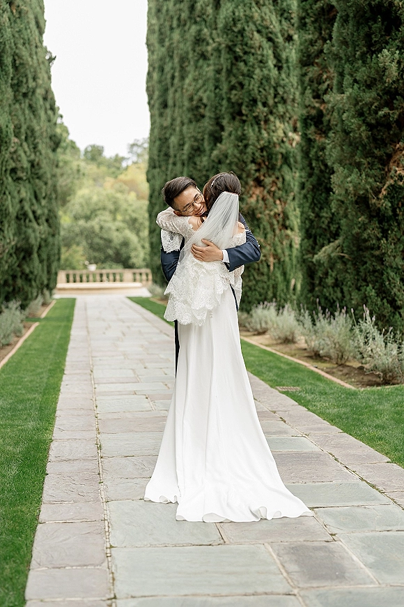 Couple portrait of bride and groom hug on a stone garden pathway, her lace veil and long train flowing as he holds her in glasses