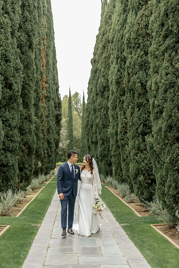 Couple portrait of bride and groom walking, bride holding bouquet with veil flowing, on a stone path along cypress trees in a garden