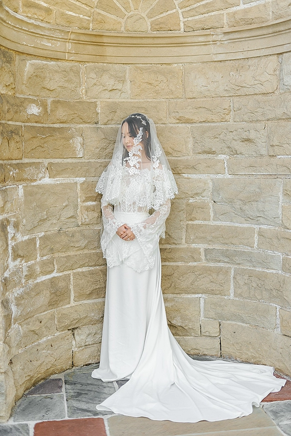 Bridal portrait of a bride in a long lace veil and lace top, hands clasped, standing by an arched stone alcove and wall