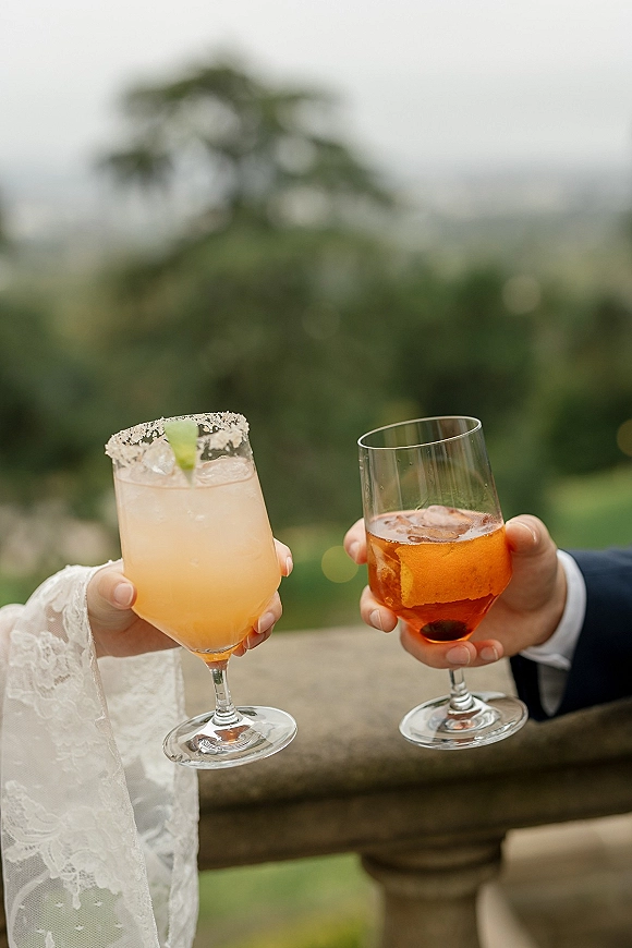 Wedding cocktail toast as bride and groom clink orange drinks with sugared rims and citrus garnish on a stone terrace with blurred greenery