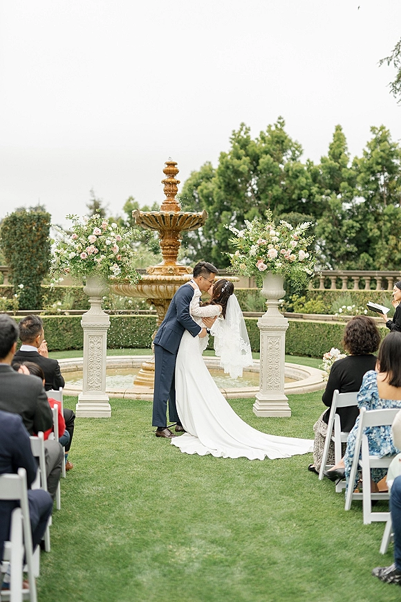 Ceremony kiss as bride in cathedral veil and long train leans into groom in navy suit on a garden ceremony aisle by a fountain