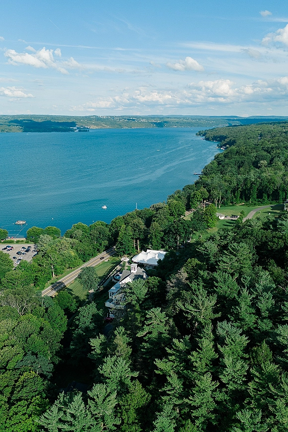 Wedding venue aerial view showing a white event tent beside a house on a lakeshore, surrounded by forest trees, boats, and clouds