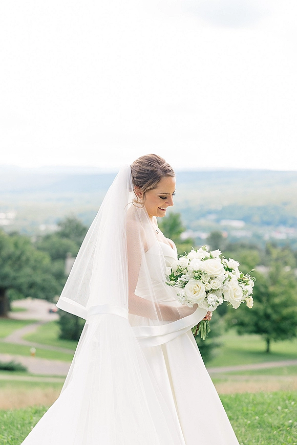 Bridal portrait of a bride holding bouquet in side profile, wearing a strapless gown and veil on a grassy hill with trees and sky