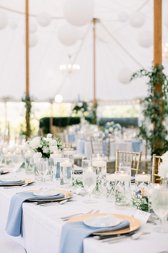 Reception tablescape with wedding table setting featuring white floral centerpieces, floating candles, blue napkins, and gold chargers under chandeliers in a white tent