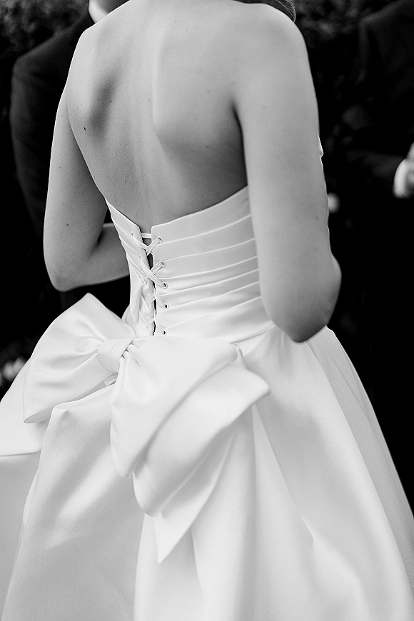 Wedding dress back with corset back wedding dress lacing in smooth satin and an oversized bow, groom suit and dark foliage behind