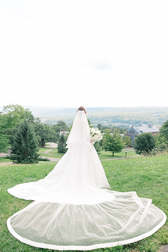 Bridal portrait of a bride from behind in a cathedral veil and long train, holding a white bouquet on a grassy hilltop under cloudy sky